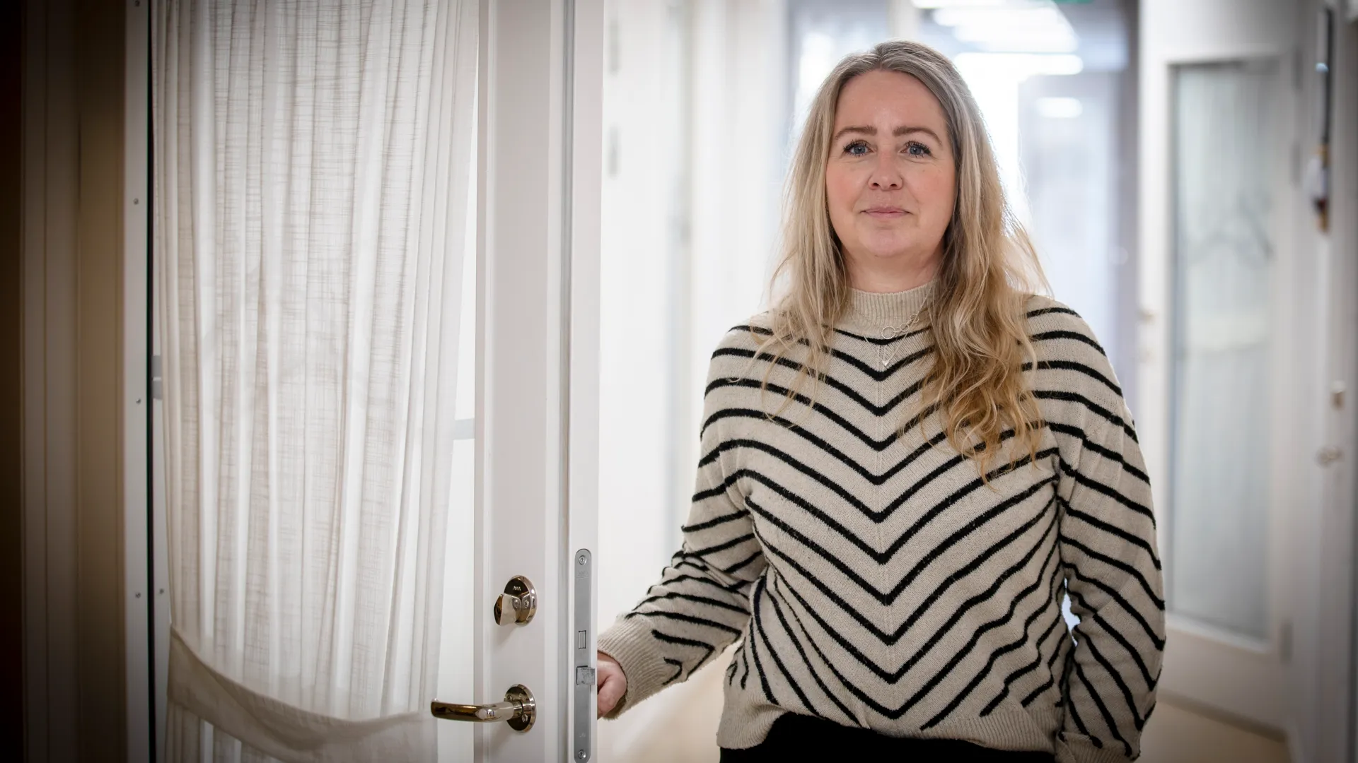 En kvinna står i en korridor och håller ena handen på ett dörrhandtag. Kvinnan kollar mot kameran.