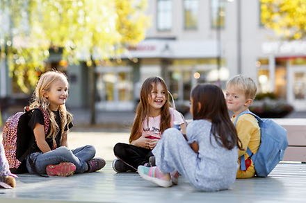 Fyra barn sitter på torget och pratar. De är glada.