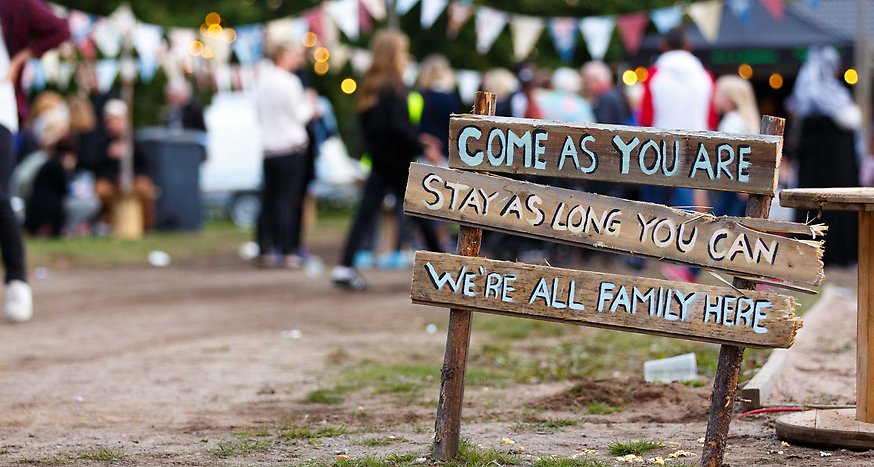 Gatufesten 2019. Människor har samlats bland vimplar, en scen och en skylt av trä som det står "Come as you are, stay as long as you can, we're all family here".