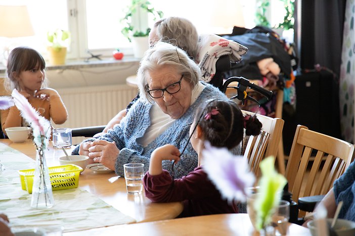Flicka sitter på stol, bredvid henne sitter två äldre personer. Hon ser in i kameran och ler.