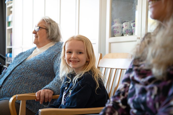 Flicka sitter på stol, ler in i kameran. Runt henne sitter två äldre personer.