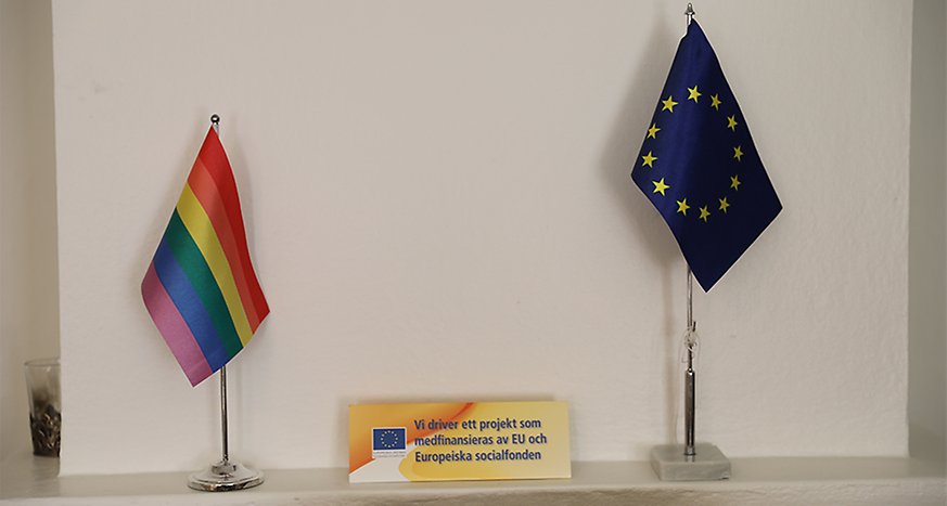 På en hylla står en regnbågsflagga, en EU-flagga och en skylt som det står att projektet medfinansieras av EU och ESF.